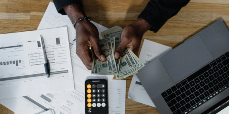 Man counting $20 bills with printed charts, laptop, and calculator app.