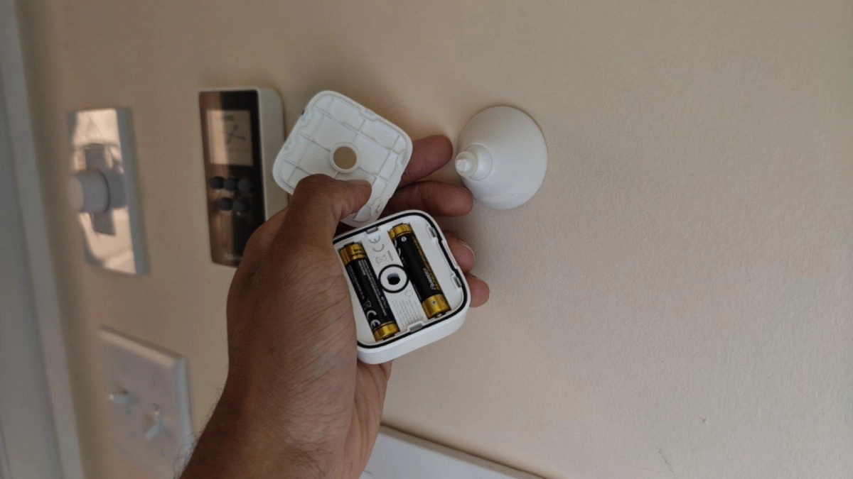 Hand holding a SwitchBot mmWave presence sensor with back cover removed and batteries visible