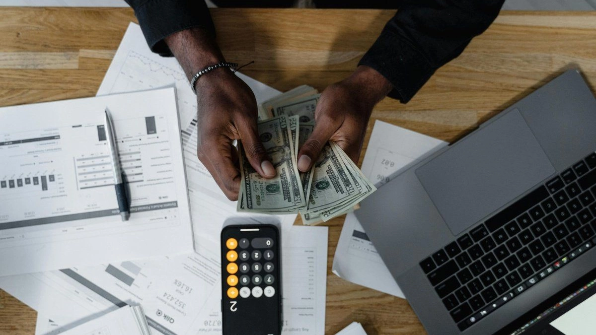 Man counting $20 bills with printed charts, laptop, and calculator app.